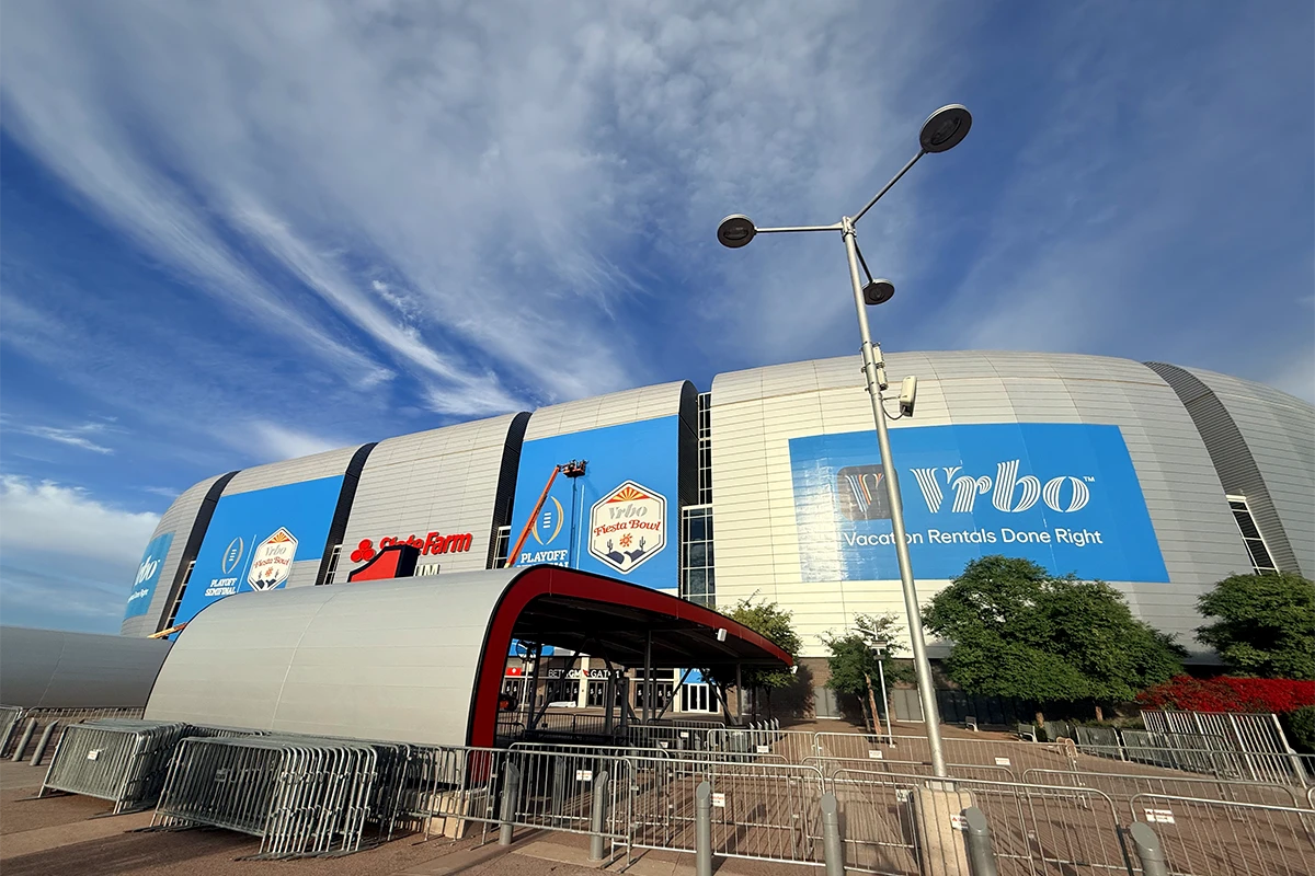 Exterior view of a large, modern football stadium with curved silver panels and oversized blue event banners, including a Vrbo sign and a Fiesta Bowl playoff logo, under a bright sky with wispy clouds. Metal crowd barriers and a covered entry structure appear in the foreground.