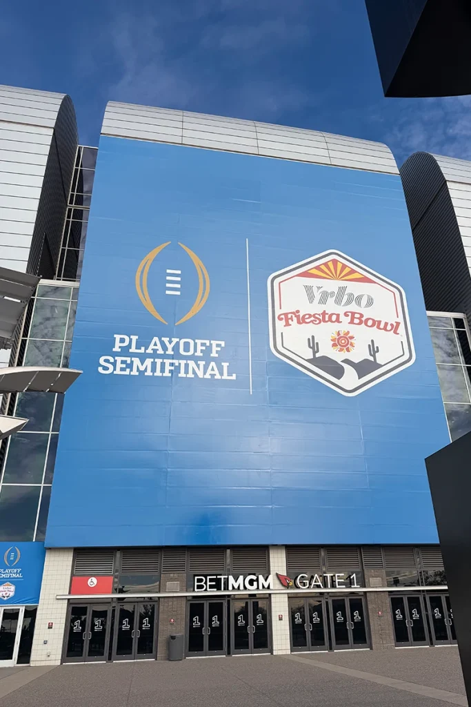 Front-facing view of a stadium entrance with a massive blue wall graphic displaying the College Football Playoff Semifinal logo alongside the Vrbo Fiesta Bowl emblem. Below the signage, multiple glass entry doors are labeled BETMGM Gate 1, with the stadium’s curved metal exterior and blue sky above.