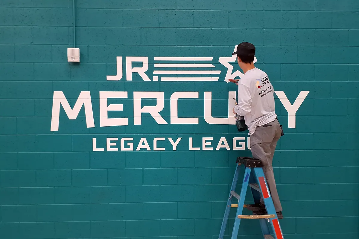 Image Craft installer applying white vinyl lettering and logos to an interior gymnasium wall for the JR. Mercury Legacy League using precision installation techniques.